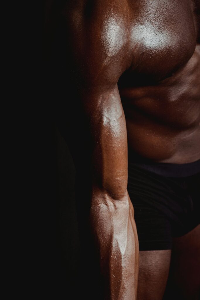 A dark-toned close-up of a shirtless man's muscular arm and torso showcasing defined muscles.